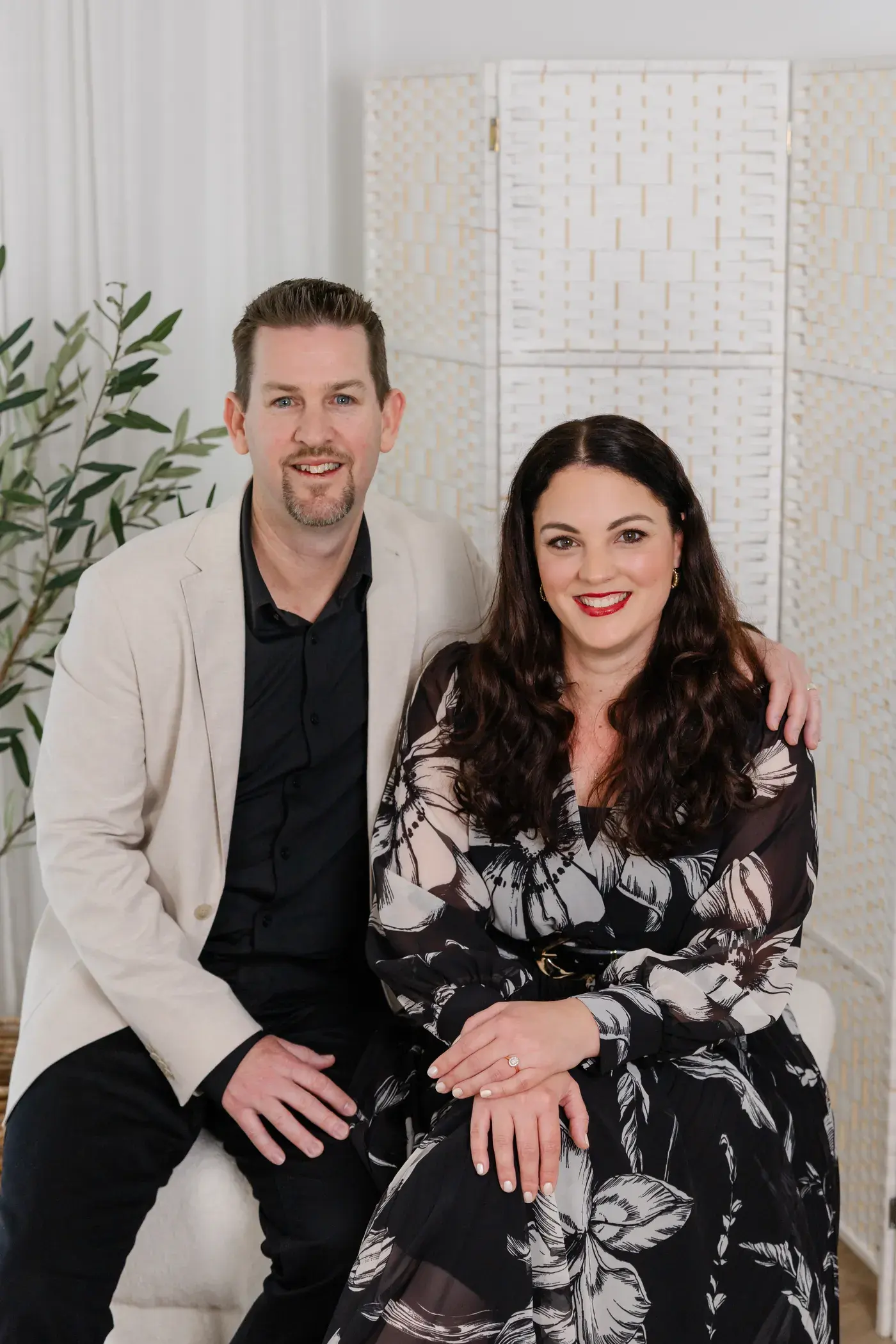 Mathew McCullagh and Shelley Walsh-McCullagh seated together and smiling, Mathew in a cream jacket with his arm around Shelley, in front of a woven white screen